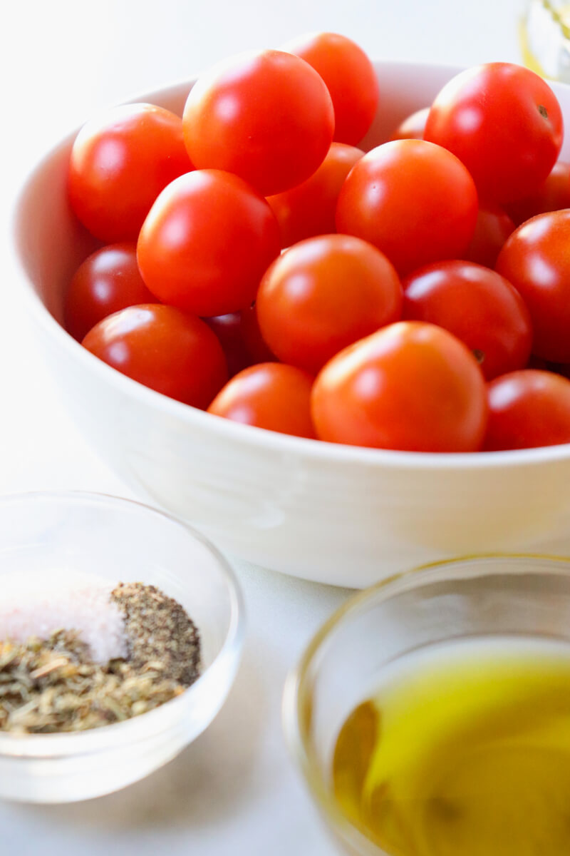 A white ceramic bowl of cherry tomatoes with smaller glass bowls of olive oil and spices in front