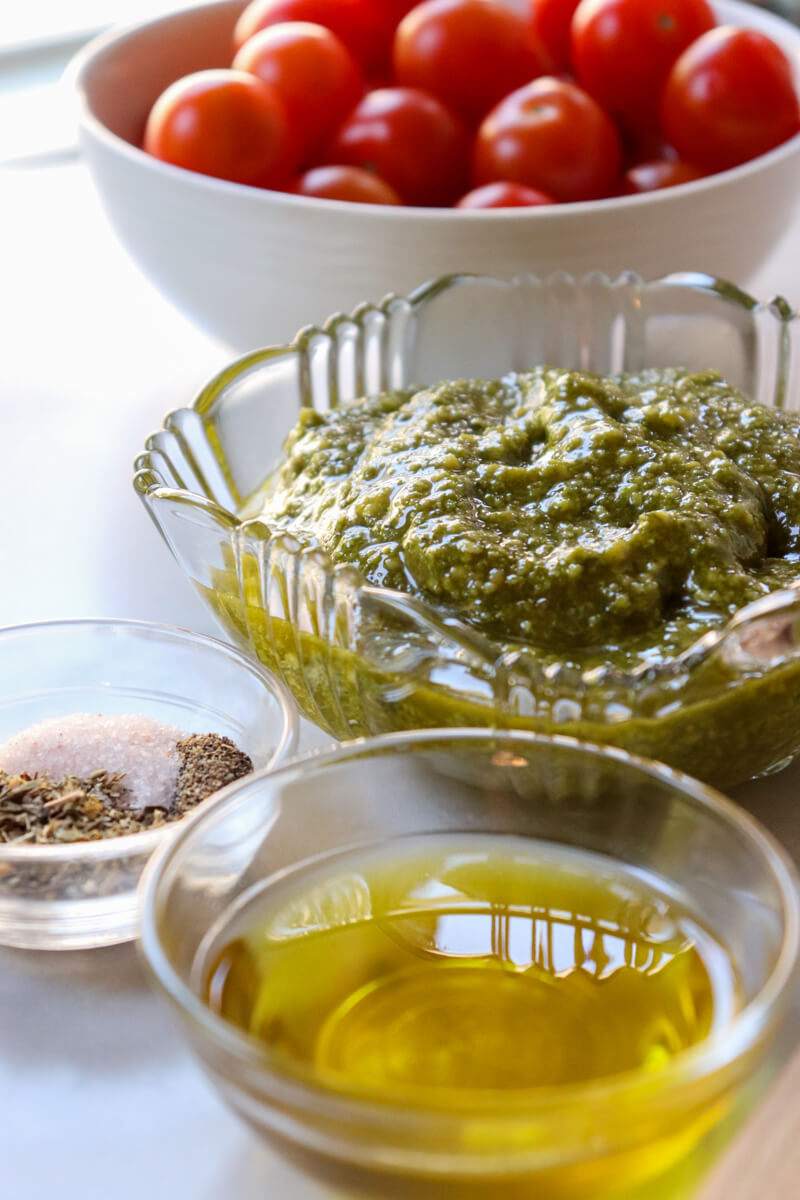 A white bowl of cherry tomatoes behind a glass beveled bowl of pesto, behind a smaller glass bowl of olive oil